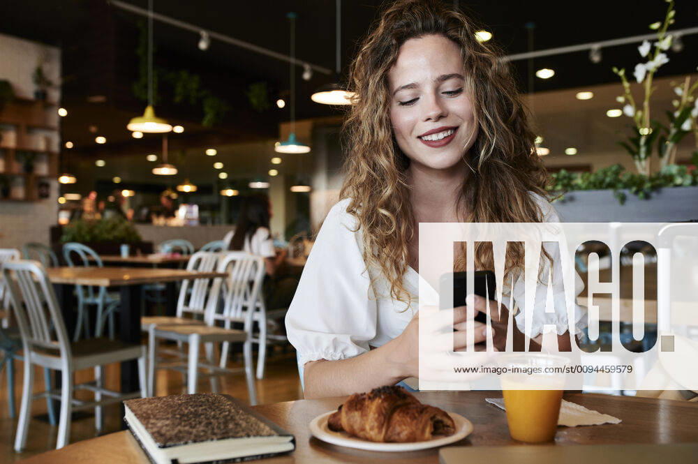 Smiling young woman using smartphone in a cafe while having breakfast model released Symbolfoto IGGF01332