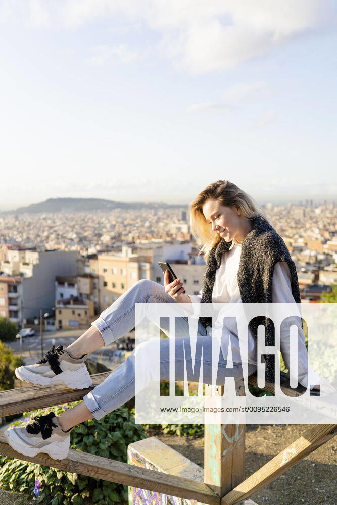 Young woman sitting on railing above the city using cell phone, Barcelona, Spain model released Symbolfoto GIOF07717