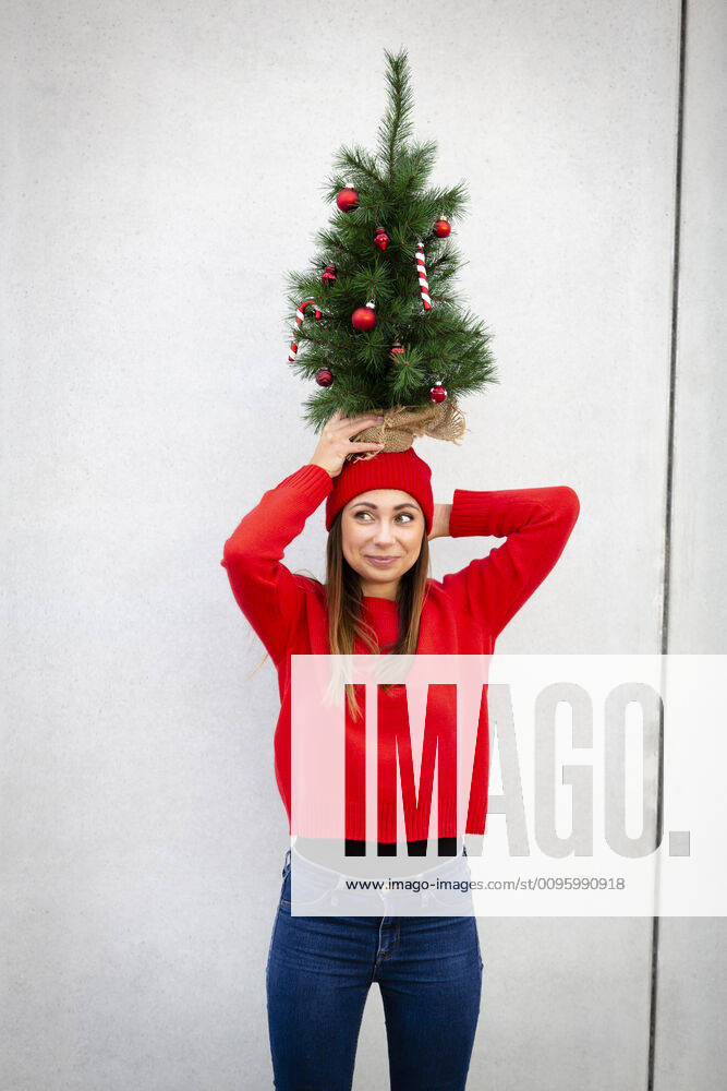 Woman wearing red pullover and wolly hat, holding artificial Christmas tree on top of her head, in front of a wall model released Symbolfoto HMEF00689