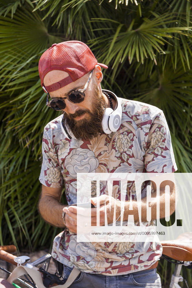 Mature man with red basecap, sunglasses and white headphones using smartphone model released Symbolfoto TCF06223