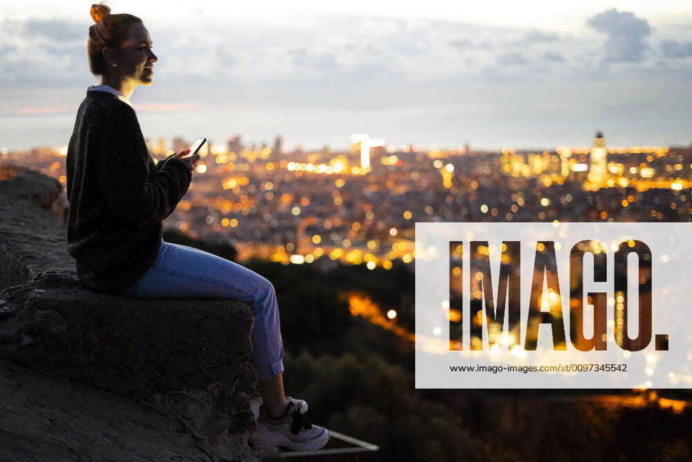 Young woman sitting on railing above the city using cell phone, Barcelona, Spain model released Symbolfoto GIOF07941