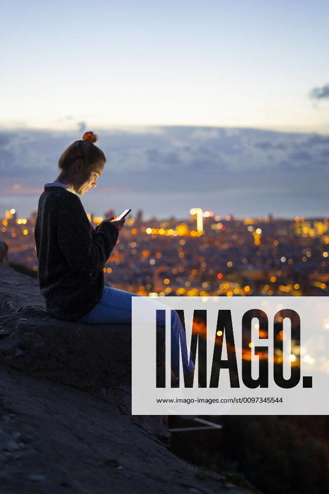 Young woman sitting on railing above the city using cell phone, Barcelona, Spain model released Symbolfoto GIOF07942