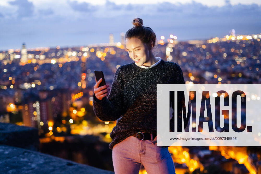 Young woman sitting on railing above the city using cell phone, Barcelona, Spain model released Symbolfoto GIOF07943
