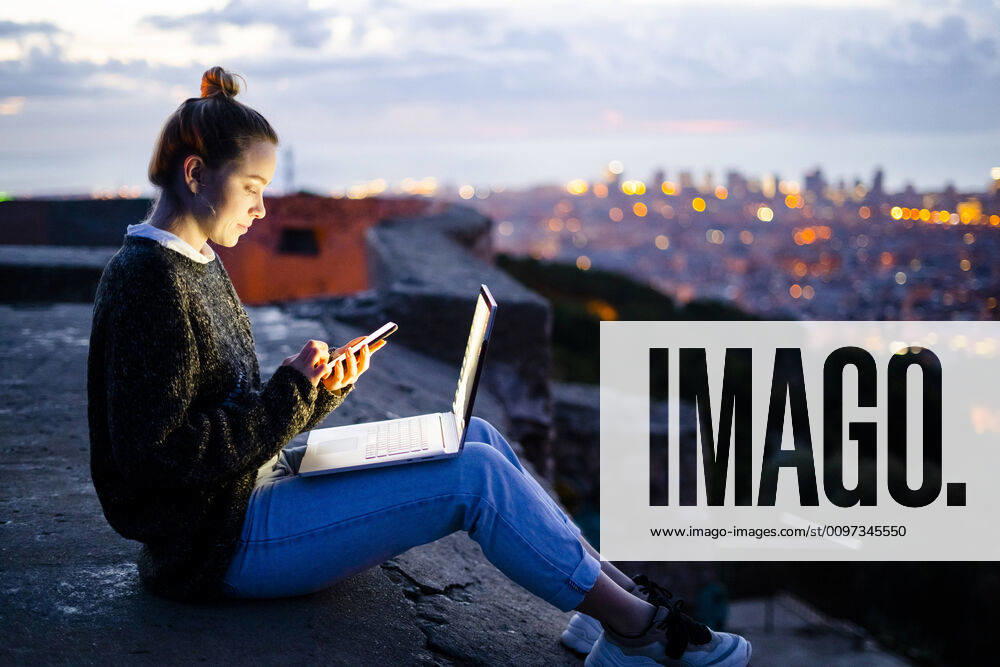 Young woman sitting on railing above the city using cell phone, Barcelona, Spain model released Symbolfoto GIOF07945