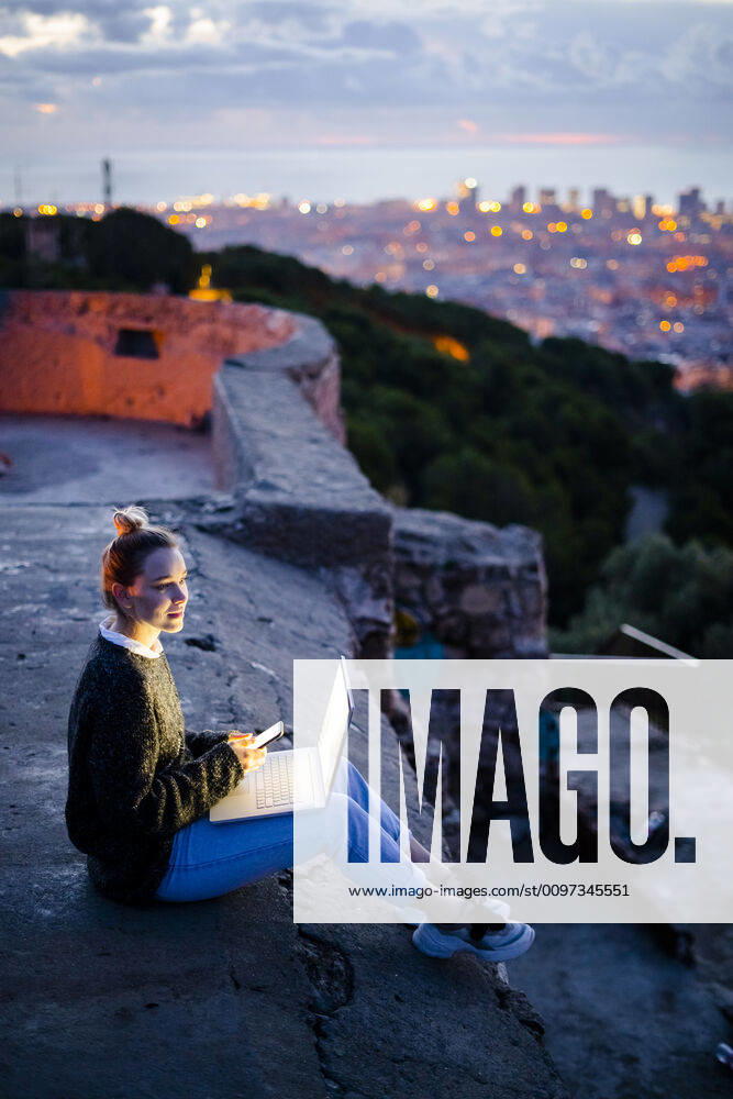 Young woman sitting on railing above the city using cell phone, Barcelona, Spain model released Symbolfoto GIOF07946