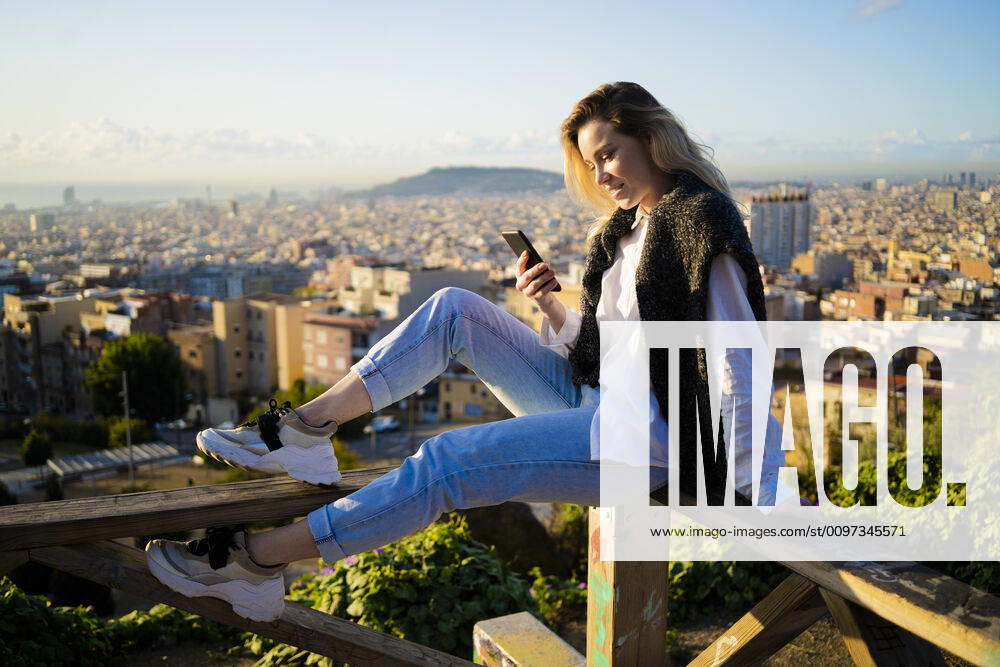 Young woman sitting on railing above the city using cell phone, Barcelona, Spain model released Symbolfoto GIOF07949