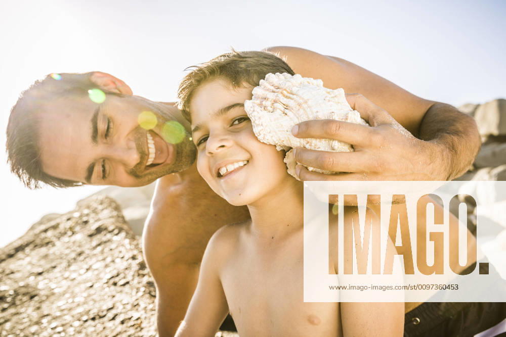 Father and son having fun on the beach and listening to the ocean in a shell model released Symbolfoto SDAHF00486