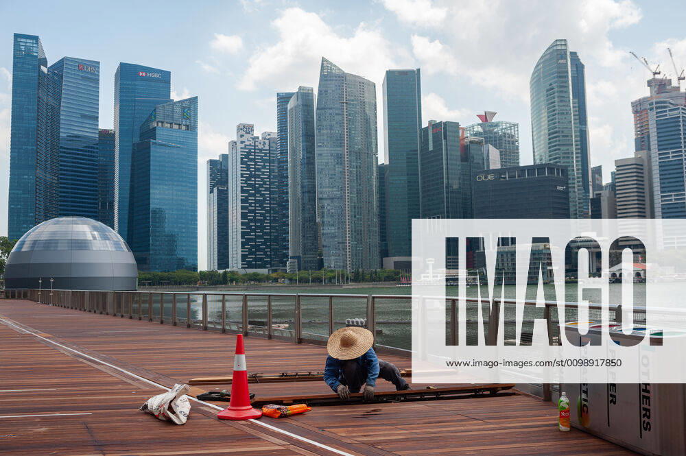 Singapur, Republik Singapur, Asien - Ein Arbeiter repariert in Marina Bay Holzplanken entlang der Uferpromenade und der Skyline des Geschaeftsviertels im Hintergrund. Seit dem Ausbruch der Corona-Pandemie ist die Zahl auslaendischer Besucher komplett zurueck gegangen. Fuer gewoehnlich wimmelt die Gegend um Marina Bay Sands von Touristen, ist jetzt aber bis auf wenige Personen total verwaist.
