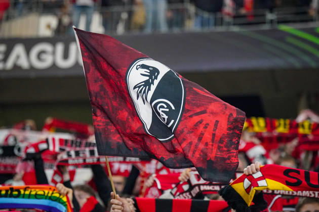 Deutschland: Fussball UEFA Conference League 2026 - 2027, 1. Spieltag - SC Freiburg vs FK Jablonec - Europa-Park-Stadion (Freiburg): Rot-schwarze Fahne mit dem Vereinswappen des SC Freiburg auf der Fantribüne im Europa-Park-Stadion beim UEFA Conference League-Spiel gegen FK Jablonec. Im Hintergrund zahlreiche Anhänger mit hochgehaltenen Fanschals, darunter ein Regenbogenschal. SC Freiburg, FK Jablonec, Fussball, Europapokal, Vereinsfahne, Schwenkfahne, Vereinslogo, Vereinswappen, Fans, Zuschauer, Fankultur, Fanblock, Heimfans, Supporter, Stimmung, Fanartikel, Regenbogen, Vielfalt, Diversität, Toleranz, Stadion. Copyright: xJOERANxSTEINSIEKx