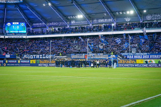 Deutschland: Fussball Bundesliga 2026 - 2027, 7. Spieltag - SC Paderborn vs Hamburger SV - Home-Deluxe-Arena (Paderborn): Heimsieg des SC Paderborn 07 gegen den Hamburger SV (HSV). Spieler, Trainer und Betreuer feiern nach Spielende gemeinsam mit den Heimfans vor der Fankurve der Home-Deluxe-Arena. Siegesfeier, Jubel, Mannschaft, Mannschaftsfoto von hinten, Schulterschluss, Fans, Ultras, Zuschauer, Tribüne, Fahnen, Schals, Fangesänge, Emotionen, Bundesliga, Fussball, Heimsieg, Sportstätte, Innenansicht, Totale, Hintertorperspektive, Tor, Fanbanner, Fankultur, Zusammenhalt. Copyright: xJOERANxSTEINSIEKx