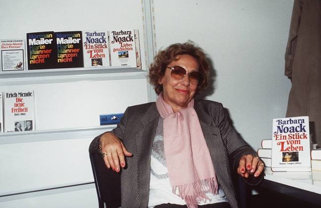 Barbara Noack (Autorin) 10 84 hör Autor Frau Literatur Buch Schriftstellerei Locken weißhaarig leger Blazer Schal Pullover sitzend aufstützend Titel Schild Ein Stück vom Leben Regal Bücherregal Tisch Brille lächelnd halb quer