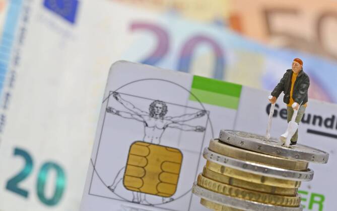 Nuernberg Symbolfoto, Themenbild Krankenversicherung, Krankenkassenbeitrag, Mann mit Gips und Gehhilfe steht auf Euro Muenzen. Im Hintergrund eine Gesundheitskarte der Barmer gek sowie Euro Banknoten. Copyright: xWuestx Eibner-Pressefotox EP_fwt