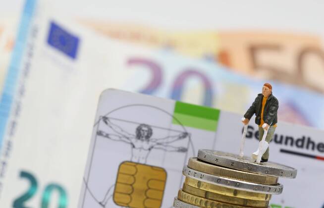 Nuernberg Symbolfoto, Themenbild Krankenversicherung, Krankenkassenbeitrag, Mann mit Gips und Gehhilfe steht auf Euro Muenzen. Im Hintergrund eine Gesundheitskarte der Barmer gek sowie Euro Banknoten. Copyright: xWuestx Eibner-Pressefotox EP_fwt