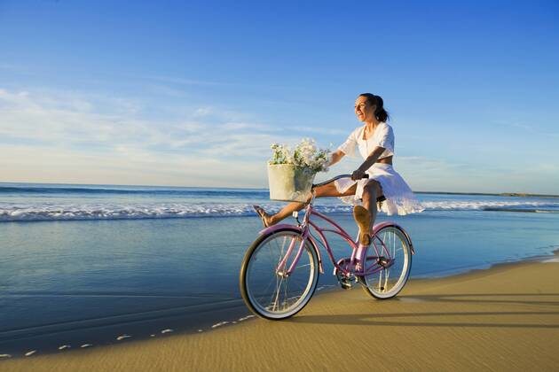 Young woman wearing a white dress riding her bike on the beach at sunset  A5