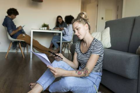 IMAGO Creative, Lifestyle, Leben in einer Wohngemeinschaft, WG Young woman sitting on floor with cell phone and papers and friends in background model released Symbolfoto property released VABF01780