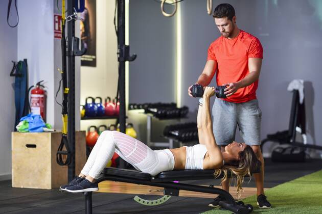Personal trainer assisting client with weight training, lifting dumbells, lying on bench model released Symbolfoto property released  JSMF00493