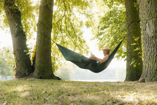 Senior man wearing straw hat relaxing in hammock at lakeshore reading book model released Symbolfoto property released GUSF01786