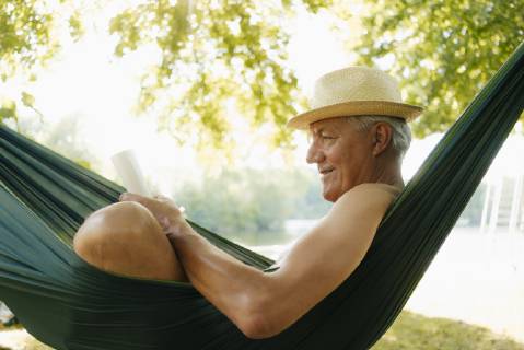 Senior man wearing straw hat relaxing in hammock at lakeshore reading book model released Symbolfoto property released GUSF01788