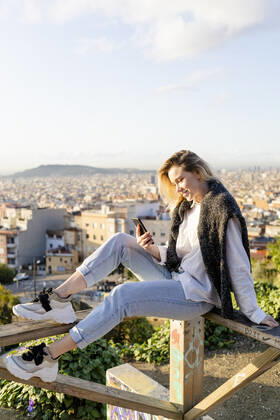 Young woman sitting on railing above the city using cell phone, Barcelona, Spain model released Symbolfoto GIOF07717