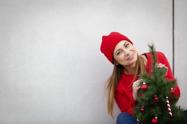 Woman wearing red pullover and wolly hat, holding artificial Christmas tree in front of a wall model released Symbolfoto HMEF00688