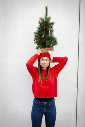 Woman wearing red pullover and wolly hat, holding artificial Christmas tree on top of her head, in front of a wall model released Symbolfoto HMEF00689