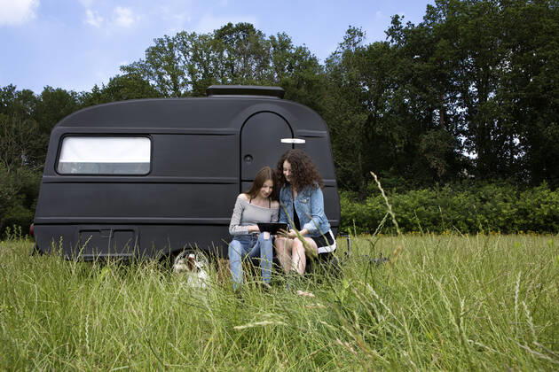 Two best friends sitting on a meadow in front of black caravan using digital tablet model released Symbolfoto property released  PSTF00489