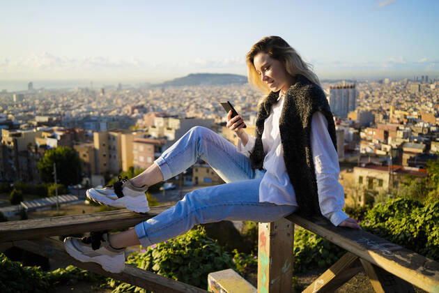Young woman sitting on railing above the city using cell phone, Barcelona, Spain model released Symbolfoto GIOF07949
