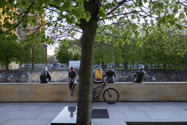 DEU, Berlin, 2020-04-18: Ein Radfahrer mit Fahrradhelm und Rucksack sitzt auf einer Mauer zwischen vier Aktfiguren der Skulptur DREI MÄDCHEN UND EIN KNABE von Wilfried Fitzenreiter, Die Skulpturen tragen Atemschutzmasken zum Schutz vor dem Coronavirus, Coronakrise, kurios, Stadtansicht, Bildhauerei, News, KBdig Foto: AKUD Lars Reimann, es gelten die AGB des Fotografen: www.akud.com