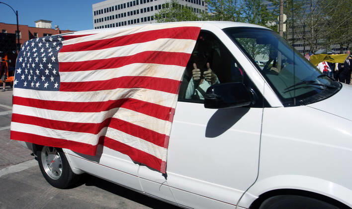 A van with a large American flag, sounds its horn, driving by the St. Louis County Government Center, during a drive-by protest in Clayton, Missouri on Tuesday, April 21, 2020. The people driving around the block plus those with signs, cheering on the sidewalks are protesting the fact that Missouri businesses are closed due to the coronavirus pandemic.  SLP2020042108 BILLxGREENBLATT