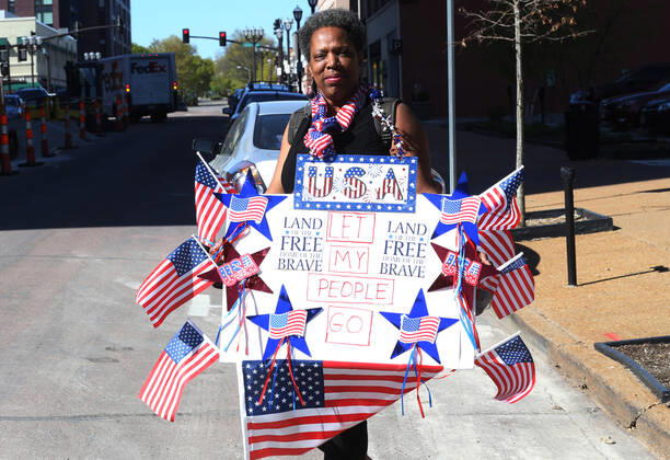 A woman with American flags crosses the street for a protest, while others drive by the St. Louis County Government Center, during a drive-by protest in Clayton, Missouri on Tuesday, April 21, 2020. The people driving around the block plus those with signs, cheering on the sidewalks are protesting the fact that Missouri businesses are closed due to the coronavirus pandemic.  SLP2020042120 BILLxGREENBLATT