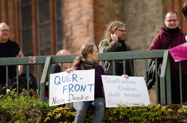 Lockdown beenden sofort! - Demonstration auf dem Südermarkt von Flensburg Die Kommunikationsstelle Demokratischer Widerstand veranstaltet eine Demonstration am 25.04.2020 in Flensburg auf dem Südermarkt, Innenstadt; hier: Gegendemonstrantin, Hanna Poddig sitzt auf der Südermarkt-Plattform, hält Transparente vor mit der Aufschrift Querfront nein Danke und Skpetisches Denken statt kruder Verschwörung