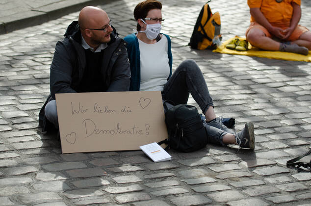 Lockdown beenden sofort! - Demonstration auf dem Südermarkt von Flensburg Die Kommunikationsstelle Demokratischer Widerstand veranstaltet eine Demonstration am 25.04.2020 in Flensburg auf dem Südermarkt, Innenstadt; zwei Demonstranten sizten auf dem Boden, vor sich Transparent mit der Aufschrift Wir lieben Demokratie , davor Lektüre des Grundgesetzes