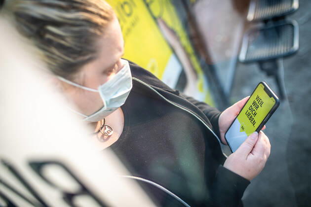 Ein Frau mit einer Mund-Nasen-Maske nutzt die BVG -App auf ihrem Smartphone an einer Bushaltestelle der BVG in Berlin. Ab Montag, den 27. April 2020 gilt eine Maskenpflicht im ÖPNV. Maskenpflicht im ÖPNV