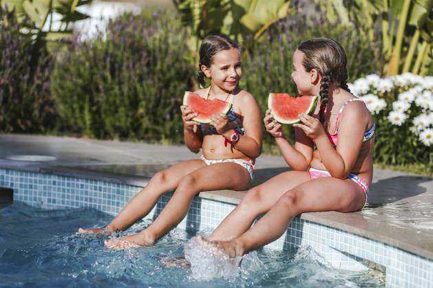 Happy twin girls sitting with watermelon slice at poolside on sunny day model released Symbolfoto property released LJF01574