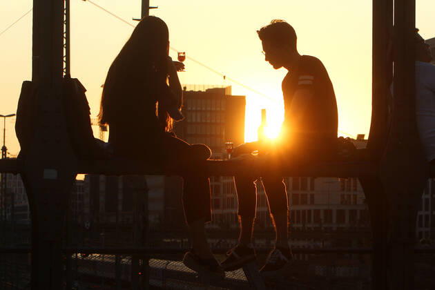 Ein Paar sitzt im Gegenlicht der untergehenden Sonne auf einem Träger der Hackerbrücke in München und unterhält sich, Bayern, Deutschland.