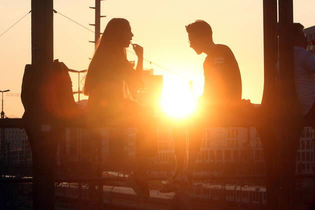 Ein Paar sitzt im Gegenlicht der untergehenden Sonne auf einem Träger der Hackerbrücke in München und unterhält sich, Bayern, Deutschland.