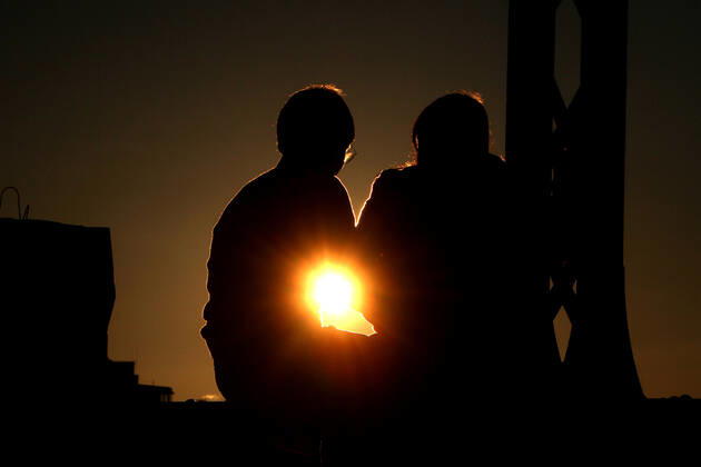 Ein Paar sitzt im Gegenlicht der untergehenden Sonne auf einem Träger der Hackerbrücke in München und unterhält sich, Bayern, Deutschland.