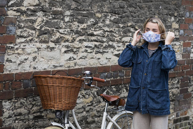 Young blond woman standing next to bicycle with basket, putting on face mask. United Kingdom,Young blond woman standing next to bicycle with basket, putting on face mask. Young blond woman standing next to bicycle with basket, putting on face mask.