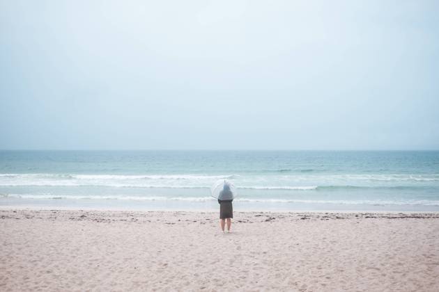 IMAGO Creative: Ende des Sommers Back view of unrecognizable traveler in raincoat with transparent umbrella walking on empty sandy beach towards waving sea in cloudy day Copyright: xJavierxBravox Model Released