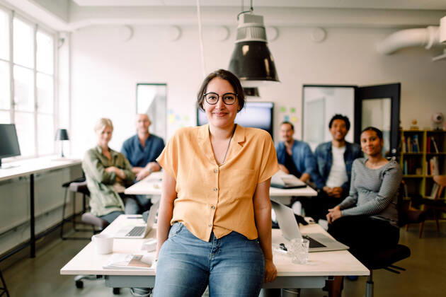 Portrait of smiling female professional leaning on table against executives in modern office , model released, property released Copyright: xMaskotx , MA73546