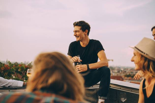 IMAGO Kreativ: Urbane Kultur - Sommer auf den Dächern Smiling man looking away while enjoying beer with friends at rooftop party against sky , model released, property released Copyright: xMaskotx , MA75644