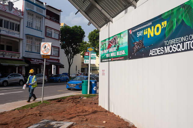 02.12.2020, Singapur, Republik Singapur, Asien - Ein Arbeiter geht waehrend der anhaltenden Coronapandemie (Covid-19) im Stadtteil Tanjong Pagar an einer Baustelle vorbei, an deren Bauzaun Transparente vor der Gefahr einer Ausbreitung und Ansteckung mit Denguefieber durch Moskitos warnen, da diese stehendes Wasser wie es auf Baustellen vorkommt, als moegliche Brutstaetten nutzen.