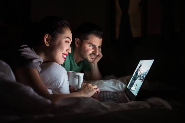 Smiling Asian female with cup of hot drink lying on bed near content boyfriend in casual wear while watching film on netbook at home Copyright: xJuanxAlbertoxRuizx Model Released
