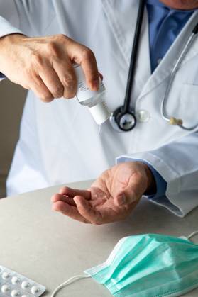 Male therapist in medical gown sitting at table in hospital and sanitizing hands with antiseptic while preparing for treatment of patients during coronavirus outbreak Copyright: xPabloxGarciax Model Released