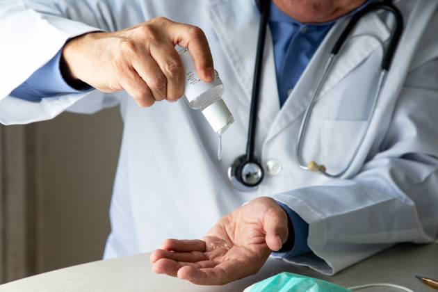 Male therapist in medical gown sitting at table in hospital and sanitizing hands with antiseptic while preparing for treatment of patients during coronavirus outbreak Copyright: xPabloxGarciax Model Released