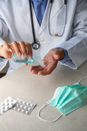 Male therapist in medical gown sitting at table in hospital and sanitizing hands with antiseptic while preparing for treatment of patients during coronavirus outbreak Copyright: xPabloxGarciax Model Released