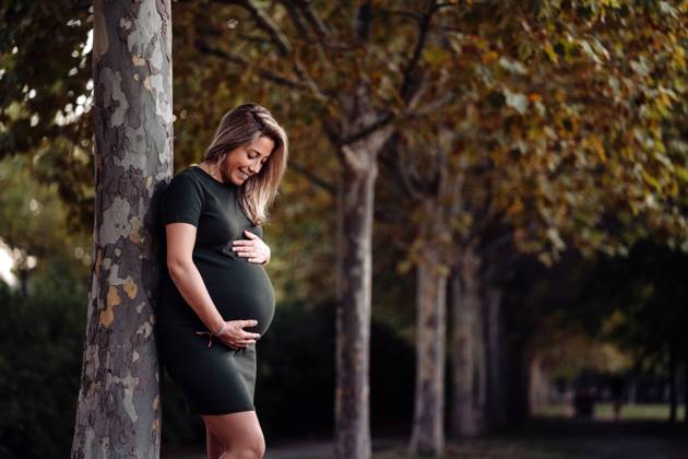 Happy pregnant woman in casual dress stroking belly while standing on green tree in sunny summer day with blue sky, Model released Copyright: xSantixMiquelx MR