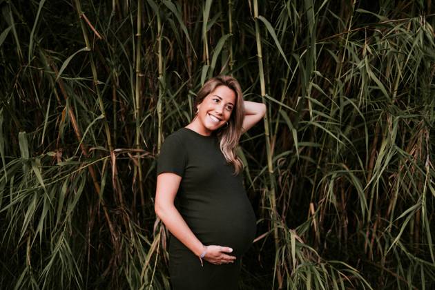 Happy pregnant woman in casual dress stroking belly while standing on green trees wall in sunny summer day with blue sky, Model released Copyright: xSantixMiquelx MR