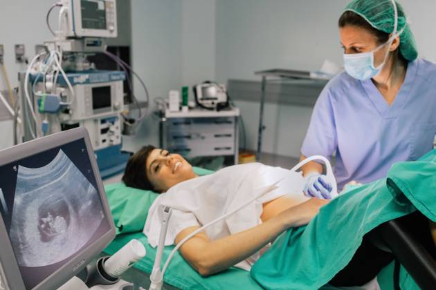 Female doctor in sterile mask and blue glove using ultrasound scanner while examining cheerful pregnant woman and looking at computer screen in hospital, Model released, Property released Copyright: xSixAndxSix MR