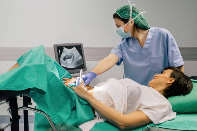 Female doctor in sterile mask and blue glove using ultrasound scanner while examining cheerful pregnant woman and looking at computer screen in hospital, Model released, Property released Copyright: xSixAndxSix MR